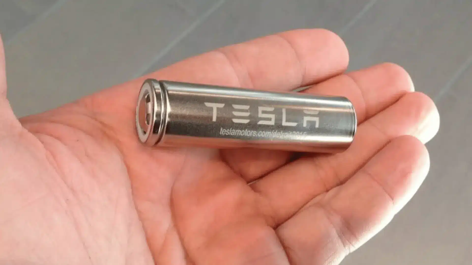a single cylindrical Tesla battery cell held in a hand, showing the branded metallic casing