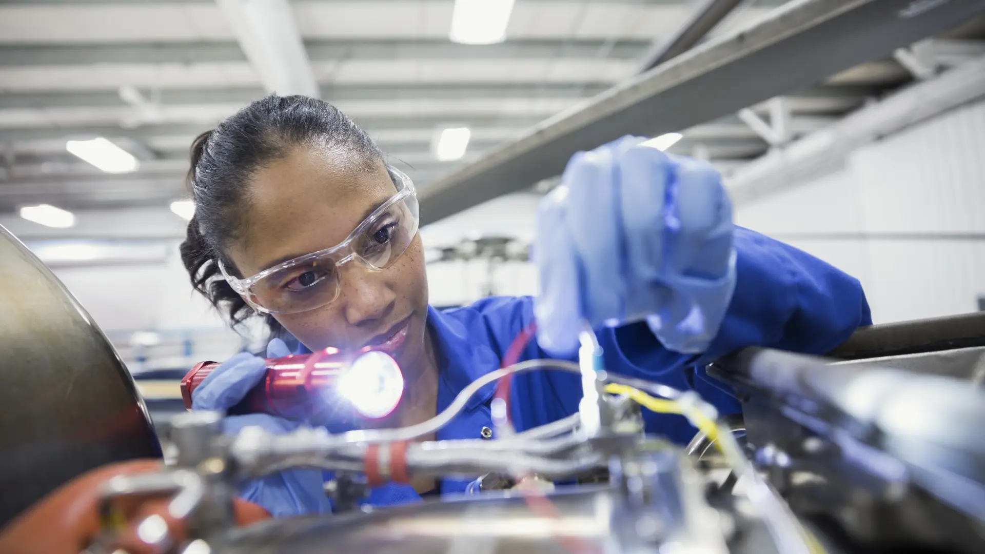 Female engineer in safety goggles using a flashlight to inspect engine wiring and components