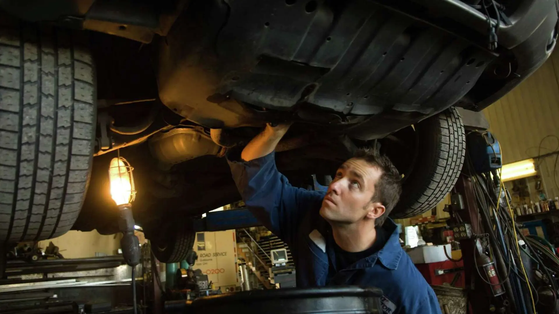 male automotive technician in coveralls inspecting the underside of a raised vehicle with a work light