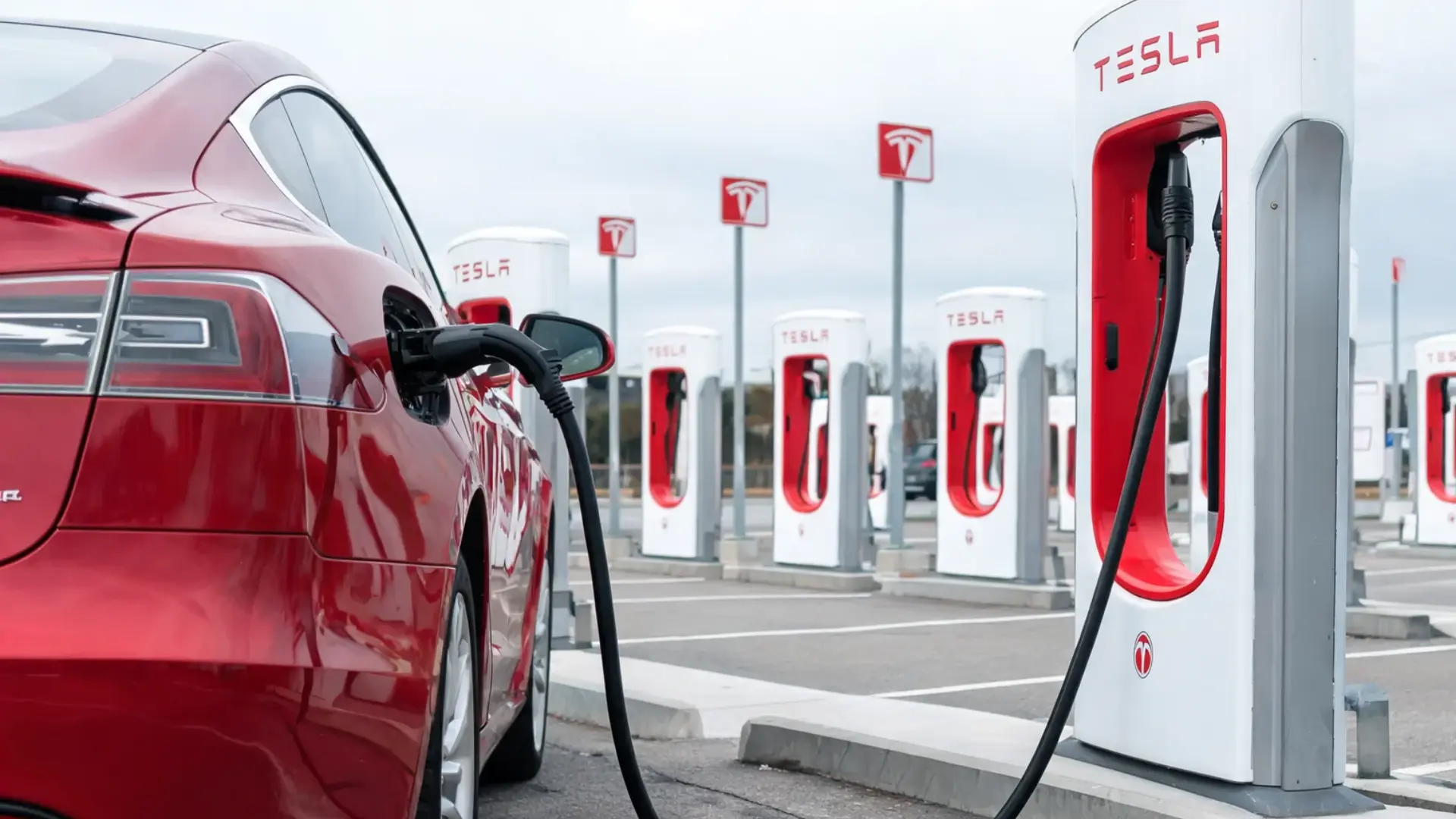 red Tesla plugged into a Supercharger station with a row of tesla chargers extending across a large lot