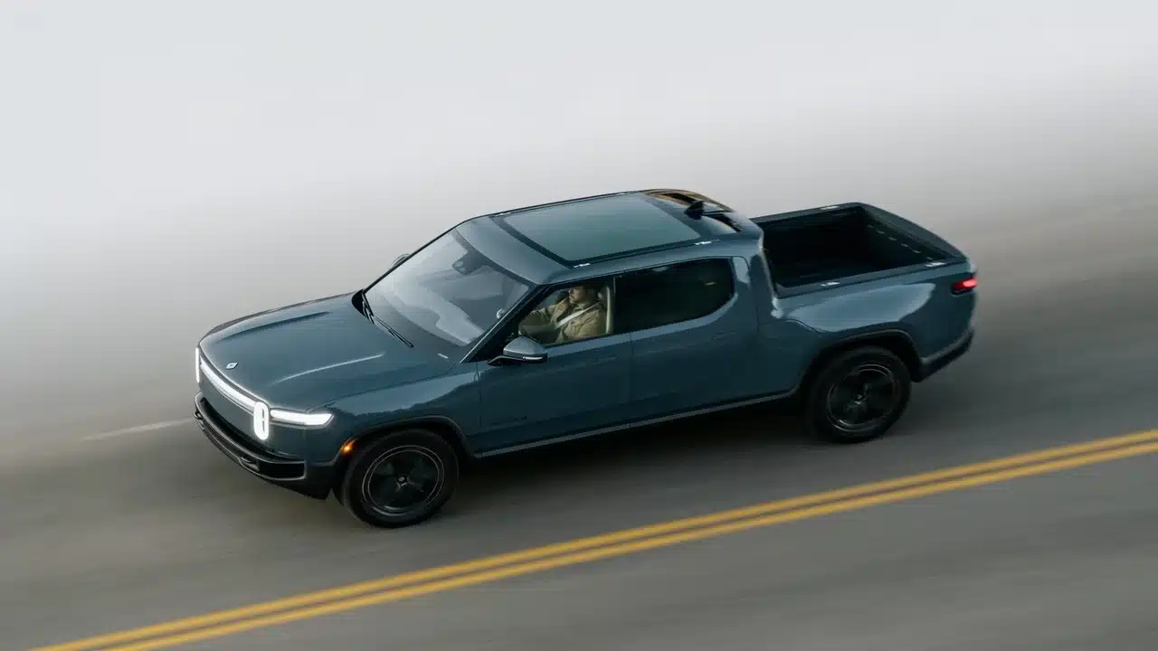 Blue Rivian R1T electric pickup driving on a foggy road with headlights on and an open truck bed.